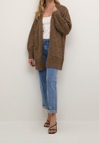 Mujer vestida con un cárdigan de punto marrón oversize sobre una blusa blanca, con jeans azul claro arremangados y tacones negros de tiras, de pie contra una pared lisa.