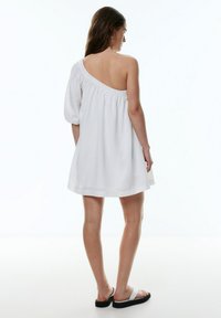 Robe blanche décolletée avec une manche à volants, coupe loose, texture en coton, longueur au genou et design uni. Portée avec des sandales blanches plates.