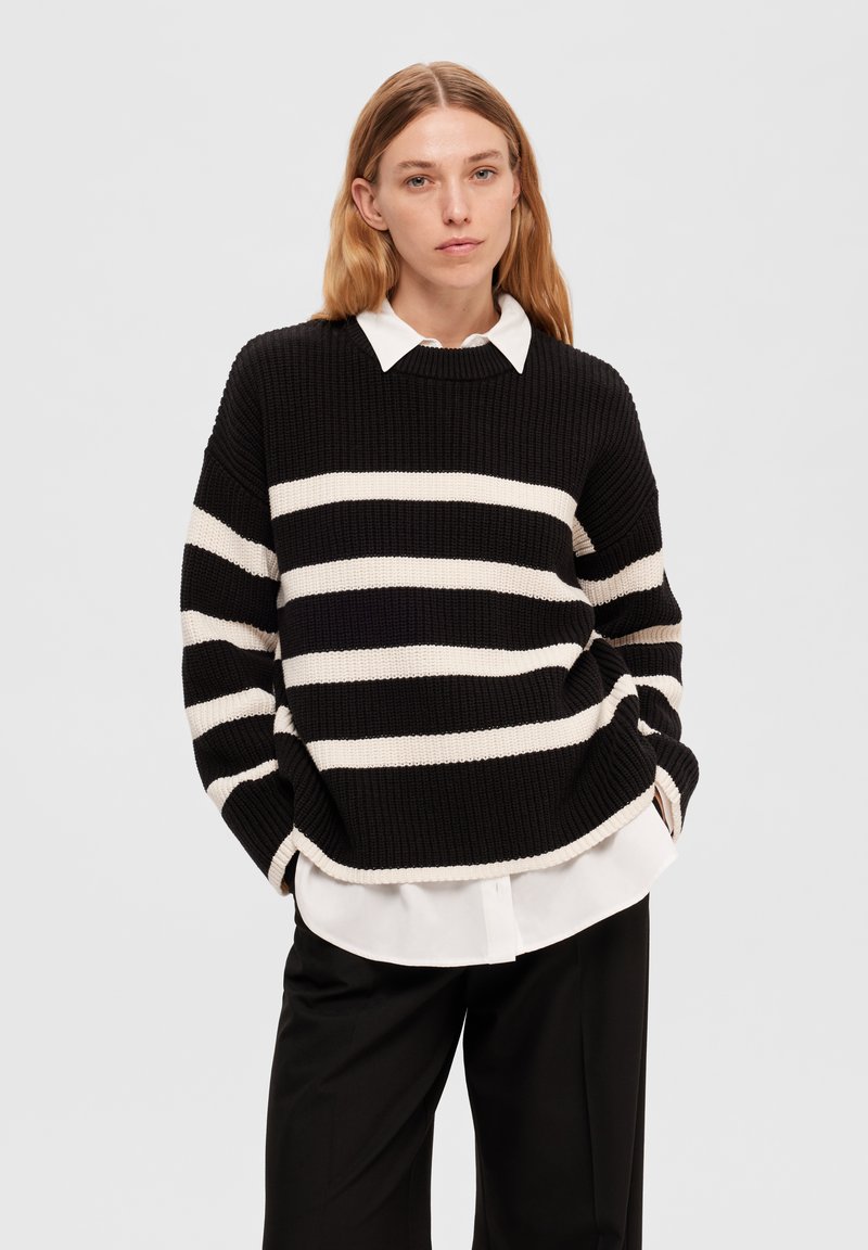Maglione a righe bianco e nero in maglia con scollatura rotonda, caratterizzato da un tessuto testurizzato e una vestibilità ampia, indossato sopra una camicia bianca con colletto.