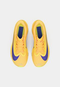 Lyse gule løpesko med mesh-materiale, lilla aksenter og teksturerte overflater. Har fremtredende snøring og polstrede såler.