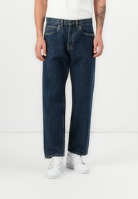 Blue denim jeans with a relaxed fit, featuring belt loops, five pockets, and contrasting stitching. Paired with white sneakers.