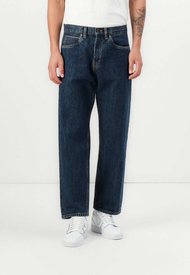 Blue denim jeans with a relaxed fit, featuring belt loops, five pockets, and contrasting stitching. Paired with white sneakers.