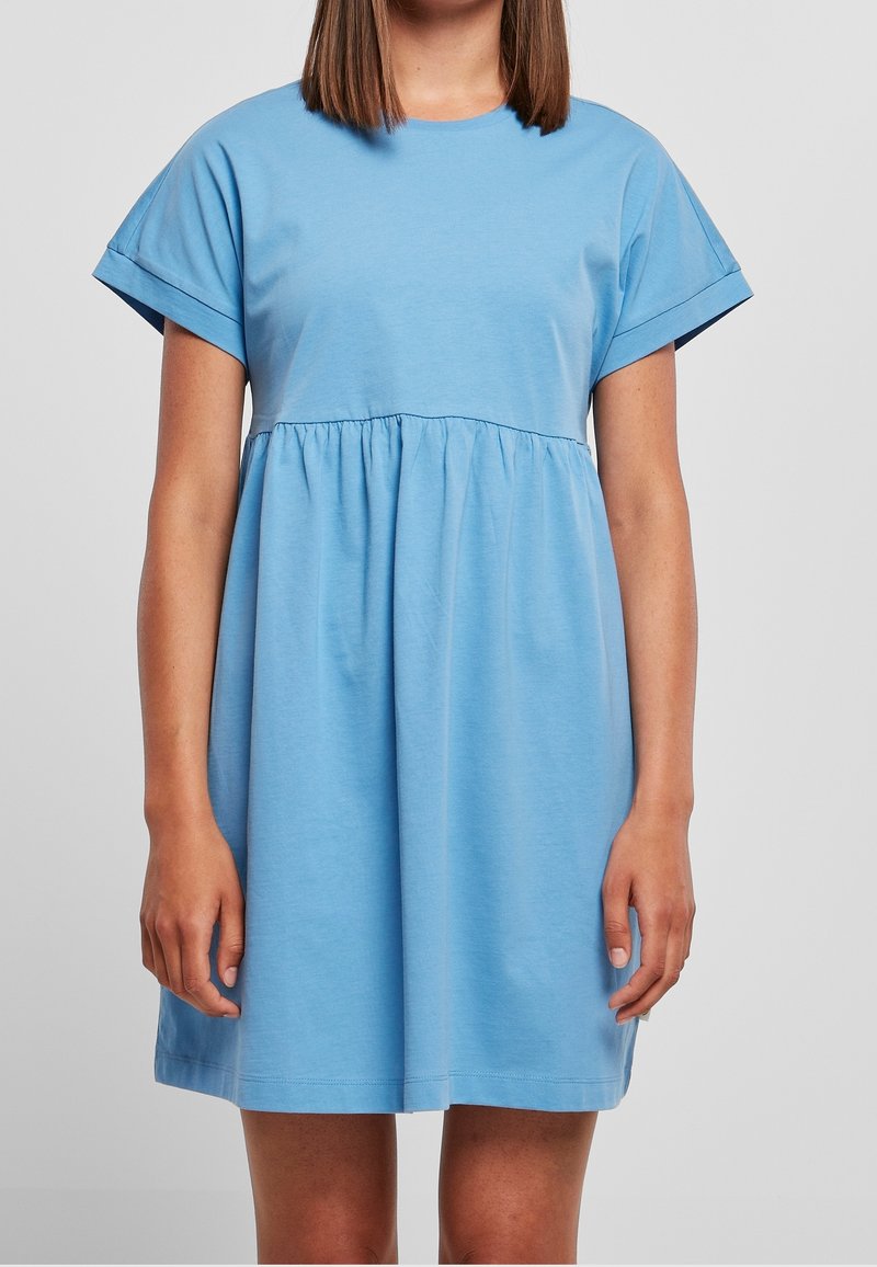 Femme portant une robe bleu clair à manches courtes, ample avec une taille froncée, debout devant un fond gris clair uni.