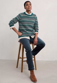 Gebreide trui in teal en marineblauw met veelkleurige geometrische patronen; gelaagd over een wit overhemd met kraag. Gecombineerd met donkere jeans en bruine laarzen.