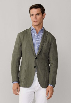Hombre con blazer verde oliva sobre camisa de rayas azules y blancas y pantalones blancos, de pie contra un fondo liso.