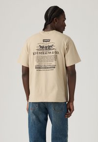 Beige bomull T-skjorte med svart grafisk trykk på baksiden, med merkets navn "Levi Strauss & Co." og dekorative detaljer.