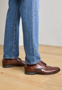Bruine leren dresschoenen met zichtbare stiksels, vetersluitingen en een zwarte rubberen zool, gecombineerd met blauwe denim jeans op een houten vloer.