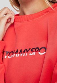 Sudadera de color coral con cuello y puños acanalados, que presenta un gran logo "TOMMY" en texto blanco y negro en la parte delantera. Textura de tela suave.