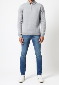 Pull gris clair en maille avec un col haut et une demi-fermeture éclair, associé à un jean bleu ajusté et des baskets blanches avec des accents verts.