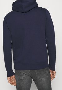 Sweat à capuche bleu marine avec une poche kangourou, des poignets et un ourlet côtelés. Tissu doux et texturé. Vue arrière montrant une silhouette lisse et la capuche.