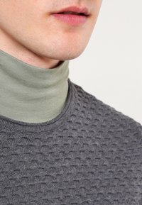 Harmaa tekstuurinen neule vaaleanvihreän poolopuseron päällä, jossa on pyöreä kaula-aukko ja 3D-kuviointi neuleen pinnalla.