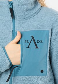 Giacca di pile blu con colletto alto, caratterizzata da una tasca a contrasto color teal decorata con un logo nero e una cerniera.