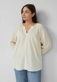 Cremfarbene, leichte Bluse mit einem strukturierten Stoff, V-Ausschnitt und lockeren, langen Ärmeln. Kombiniert mit blauen Jeans.