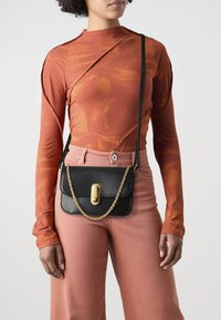 Borsa a tracolla in pelle nera con hardware dorato, tracolla a catena e chiusura a fischio. Accompagnata da un top aderente arancione a maniche lunghe.