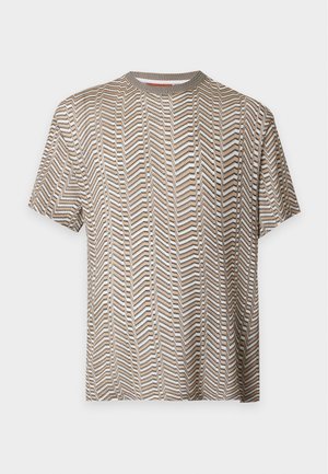 Homme debout, main dans la poche, portant une chemise à manches courtes beige et blanche à motifs, un pantalon vert olive et des chaussures noires sur fond blanc.