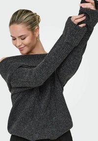 Ciemnoszary, teksturowany sweter z dekoltem na jedno ramię, długimi rękawami i ściągaczami, noszony na czarnej odzieży.