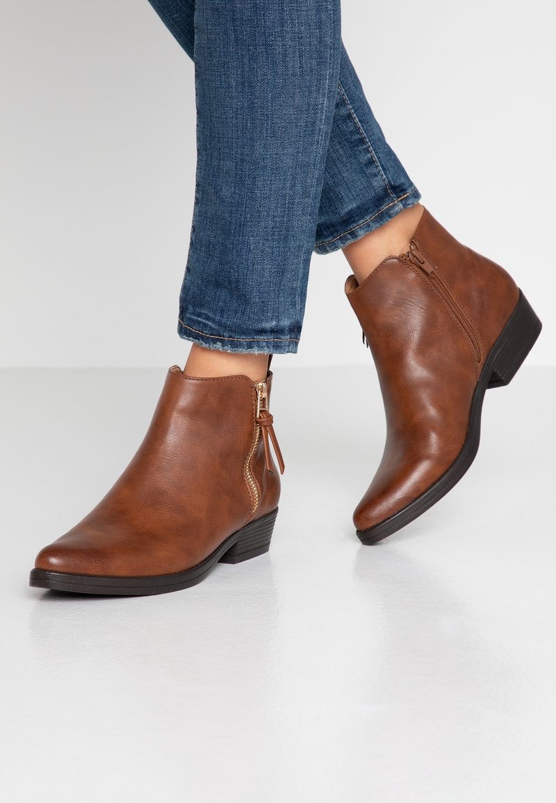 Bottes en cuir marron hauteur cheville avec des fermetures éclair latérales portées avec un jean en denim bleu sur un sol réfléchissant la lumière.