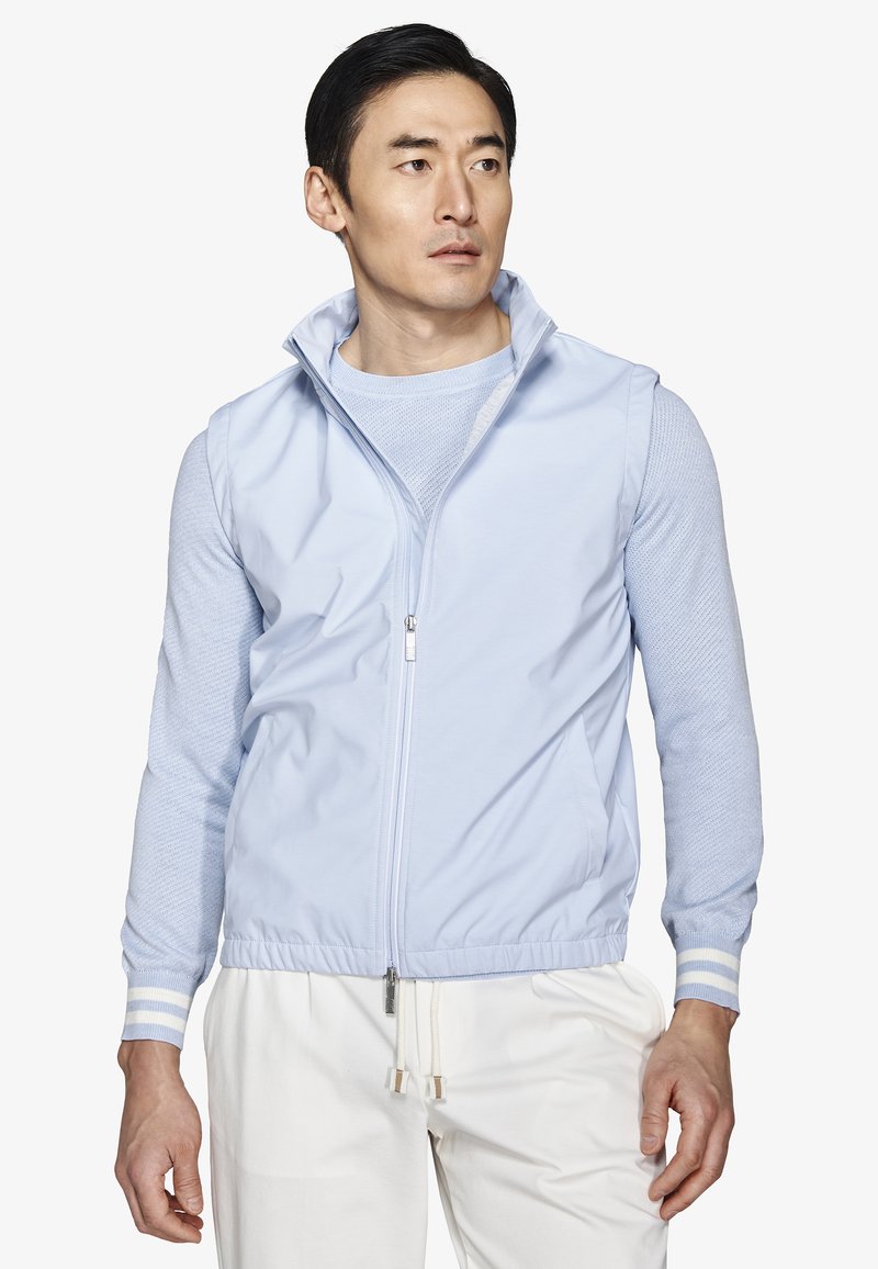 Mann trägt eine hellblaue ärmellose Reißverschlussweste über einem langärmeligen Pullover und weiße Jogginghosen mit Kordelzug, schaut nach links.