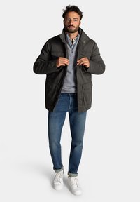 Donkergroene jas met een hoge kraag, ritssluiting en twee voorkruiken, gedragen over een grijze trui en blauwe spijkerbroek, gecombineerd met witte schoenen.