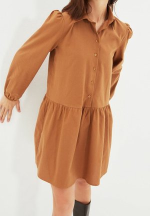 Vrouw die een bruin, lang mouwen, knoopjesjurk draagt met een geplooide taille en kraag, gecombineerd met zwarte laarzen tot aan de knie.