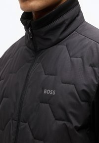 Κοντινό πλάνο ενός ατόμου που φοράει μαύρο διπλοραμμένο μπουφάν με ψηλό γιακά και ορατό λογότυπο "BOSS" στο στήθος.
