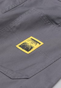 Gray fabric with a textured surface, featuring a prominent yellow rectangular patch with black graphics, sewn onto the garment.