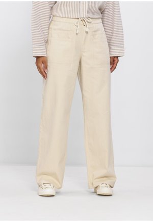 Personne portant un pantalon beige à jambes larges avec taille à cordon de serrage et poches avant, associé à des baskets blanches et une chemise beige rayée.
