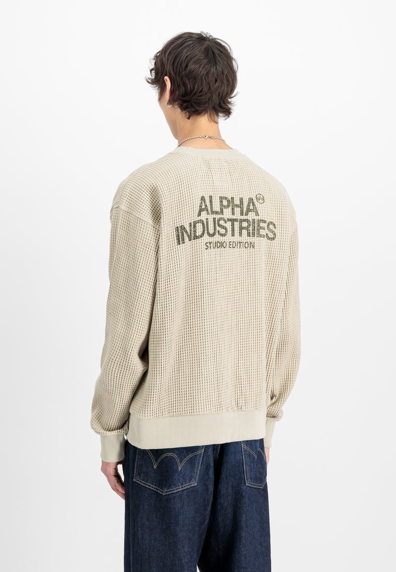 Osoba nosząca beżowy sweter o strukturze waflowej z napisem „Alpha Industries Studio Edition” na plecach, zestawiony z ciemnoniebieskimi jeansami.