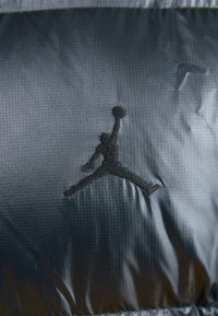 Czarna tkanina nylonowa posiada wyraźne czarne haftowane logo Jumpman, teksturowane w kratkowany wzór i o błyszczącym wykończeniu.