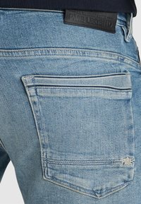 Ljusblå denimjeans har en bakficka med kontrasterande sömmar och en märkeslogotyp i svart läder nära midjan.