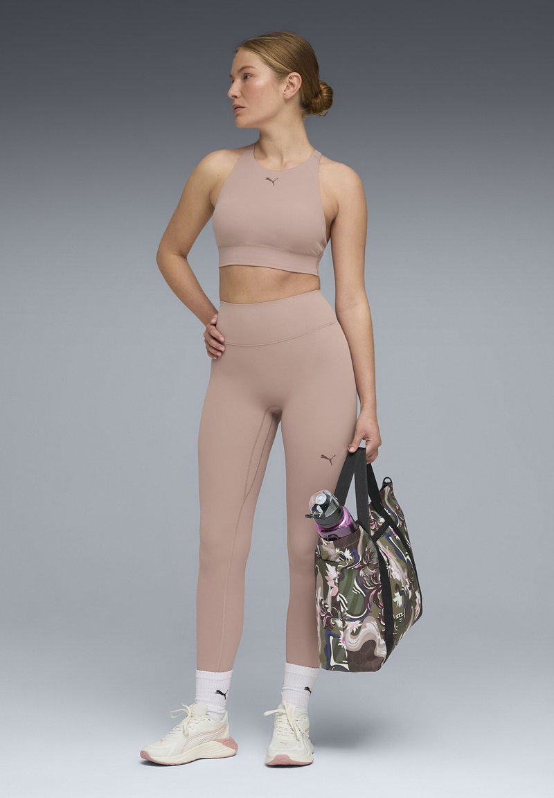 Crop top de sport rose clair et leggings taille haute. Modèle tenant un sac fourre-tout à motifs multicolores avec une bouteille d'eau. Baskets blanches.