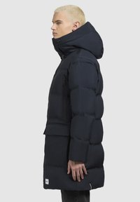 Tengerészkék pufikabát magas gallérral, cipzáras zsebekkel és megkötött derékkal. Simább textúrával és enyhén fényes felülettel rendelkezik.