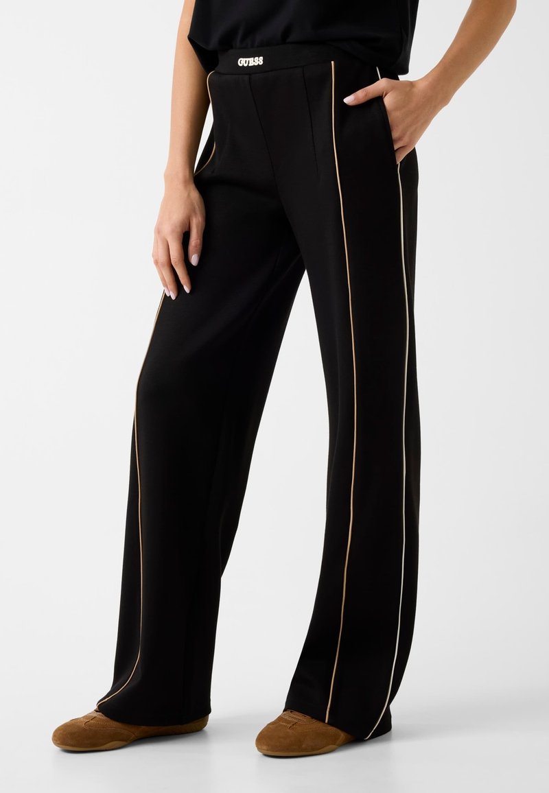 Frau trägt schwarze Hosen mit weitem Bein, beigen Seitenstreifen und braunen Wildlederschuhen, steht mit einer Hand in der Tasche und einer neben dem Körper.