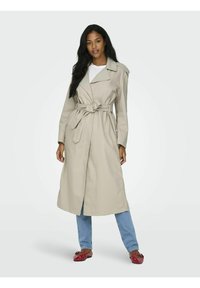 Beige trenchcoat met een brede kraag, een tailleband en zijzakken, gecombineerd met lichtblauwe jeans en rode ballerina's. Gladde, stevige stof.