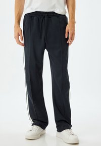 Marineblå joggerbukser lavet af blødt stof, med elastisk talje og snor samt hvide sidelister, parret med hvide sneakers.