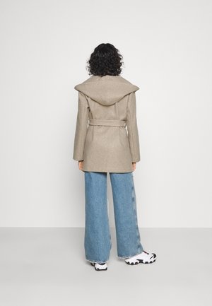 Person mit lockigem dunklem Haar, der einen beigen, mit Gürtel versehenen Mantel mit Kapuze, weite blaue Jeans und weiße Turnschuhe trägt und vor einer weißen Wand steht.