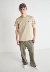 T-shirt beige in cotone con piccolo logo rosso, abbinata a pantaloni larghi verdi oliva e sneakers bianche. Cappellino in tessuto grigio tenue.