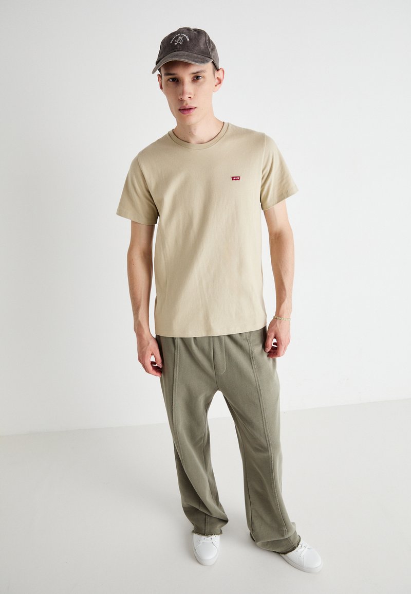 Camiseta de algodón beige con un pequeño logo rojo, combinada con pantalones sueltos verde oliva y zapatillas blancas. Gorro de tela gris apagado.