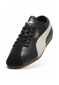 Chaussure de sport en cuir noir avec des accents en daim, présentant un panneau latéral blanc, un marquage doré, des lacets plats et une semelle en caoutchouc marron.