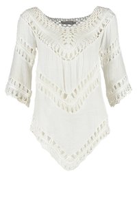 Blouse en crochet blanc avec des manches trois-quarts, un ourlet festonné et des motifs ajourés le long du décolleté, des manches et du corps.
