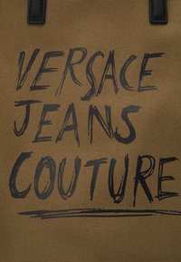 Brązowa torba płócienna z wyrazistym, czarnym napisem "VERSACE JEANS COUTURE", z czarnymi skórzanymi akcentami i teksturowaną powierzchnią tkaniny.
