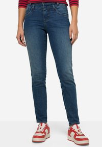 Kapeat farkut tummansinisinä, joissa on viisi taskua ja hieman haalistunut rakenne, yhdistettynä punaisiin ja valkoisiin lenkkareihin.