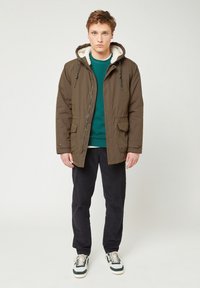 Parka marrone isolato con cappuccio foderato in pelliccia sintetica, tasche frontali e chiusura con zip, abbinato a un maglione teal e pantaloni scuri.
