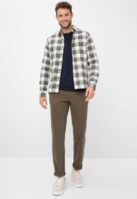 Camicia flanella a quadri verde e bianco, indossata sopra una maglietta blu navy, abbinata a pantaloni marroni e sneakers bianche.