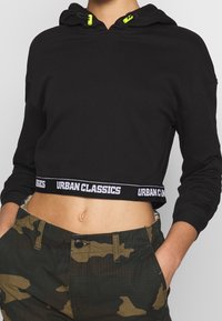 Svart kort hoodie med dragsko och ljusgula detaljer. Utformad med en kontrasterande vit band som är märkt "URBAN CLASSICS."