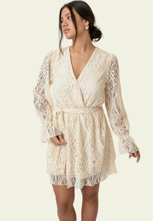 Naine, kes kannab kreemjat pitsist ümber seotud kleiti pikkade puhvis varrukatega ja kuldsete rõngaskõrvarõngastega, poseerib ühtlase heleda tausta ees.