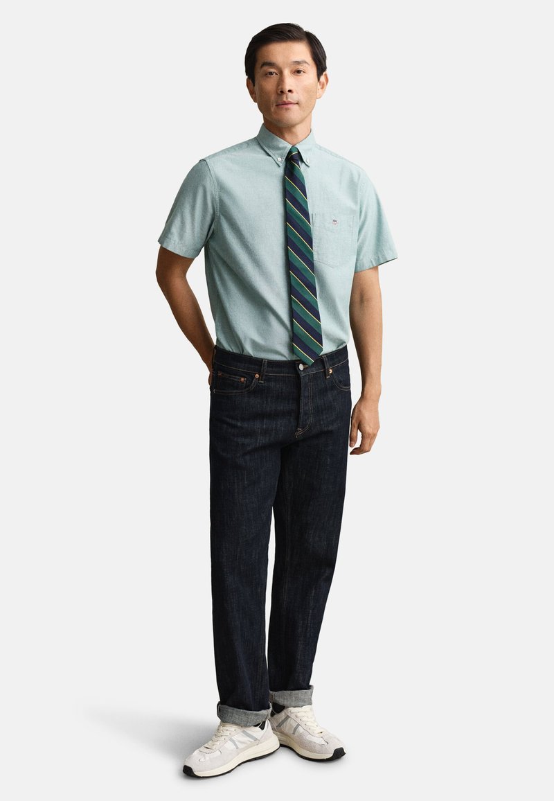 Hombre de pie con camisa de manga corta verde claro abotonada, corbata de rayas, vaqueros oscuros con puños y zapatillas blancas sobre fondo blanco.