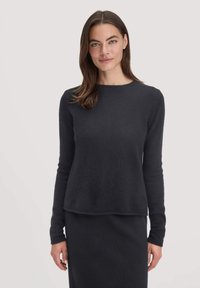 Frau mit glattem braunem Haar, die einen taillierten schwarzen Pullover mit langen Ärmeln und einen passenden Rock trägt, steht vor einem hellen Hintergrund.