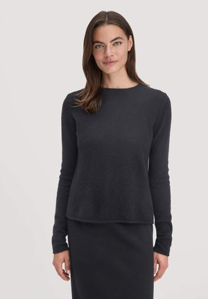Frau mit glattem braunem Haar, die einen taillierten schwarzen Pullover mit langen Ärmeln und einen passenden Rock trägt, steht vor einem hellen Hintergrund.