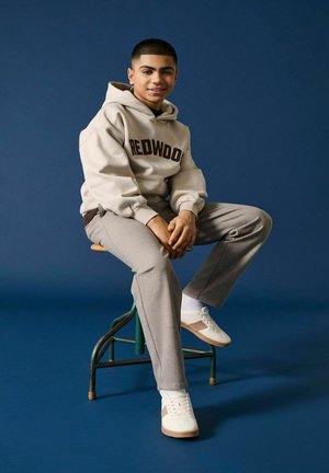 Giovane uomo seduto su uno sgabello, indossa un hoodie beige Redwood, pantaloni grigi, sneakers bianche e sorride contro uno sfondo blu scuro.